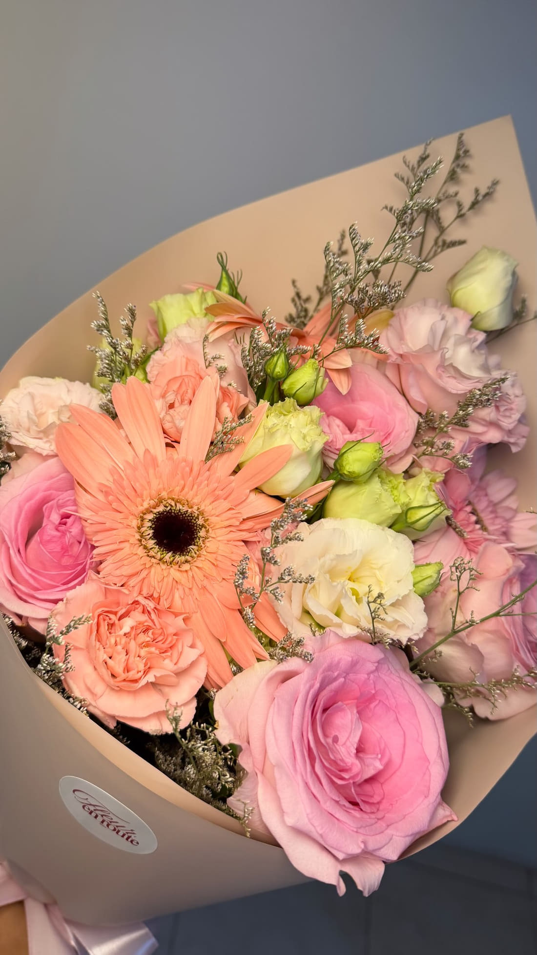 A wrapped Fleur en Route bouquet photographed from above: a peach gerbera, pink China roses, cream and pink lisianthus, peach carnations and silvery misty in kraft paper with the round Fleur en Route seal.