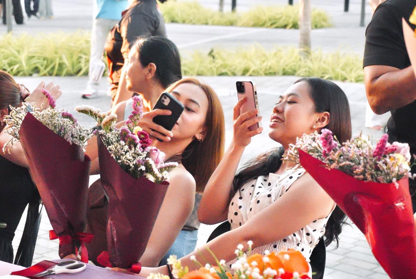 Friends photographing their wine-red wrapped bouquets at an outdoor table